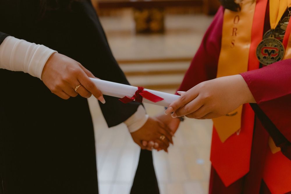 Graduate in black gown receives diploma during commencement ceremony from official in red and gold regalia.
