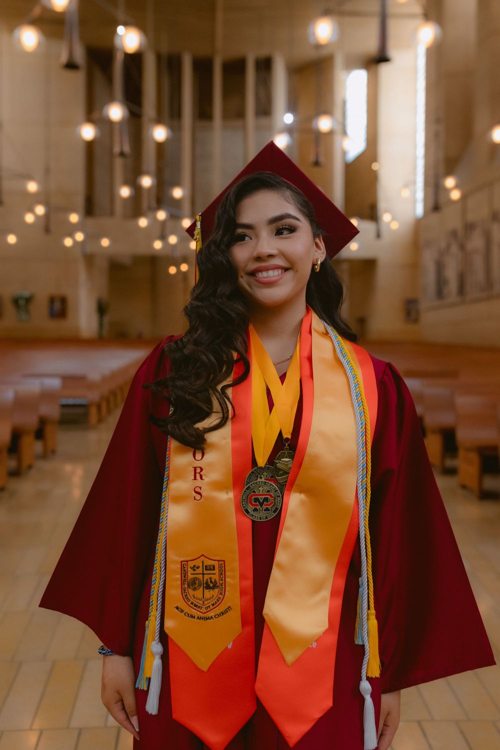 Graduate in red cap and gown with gold honors stoles standing in church sanctuary with glowing pendant lights.