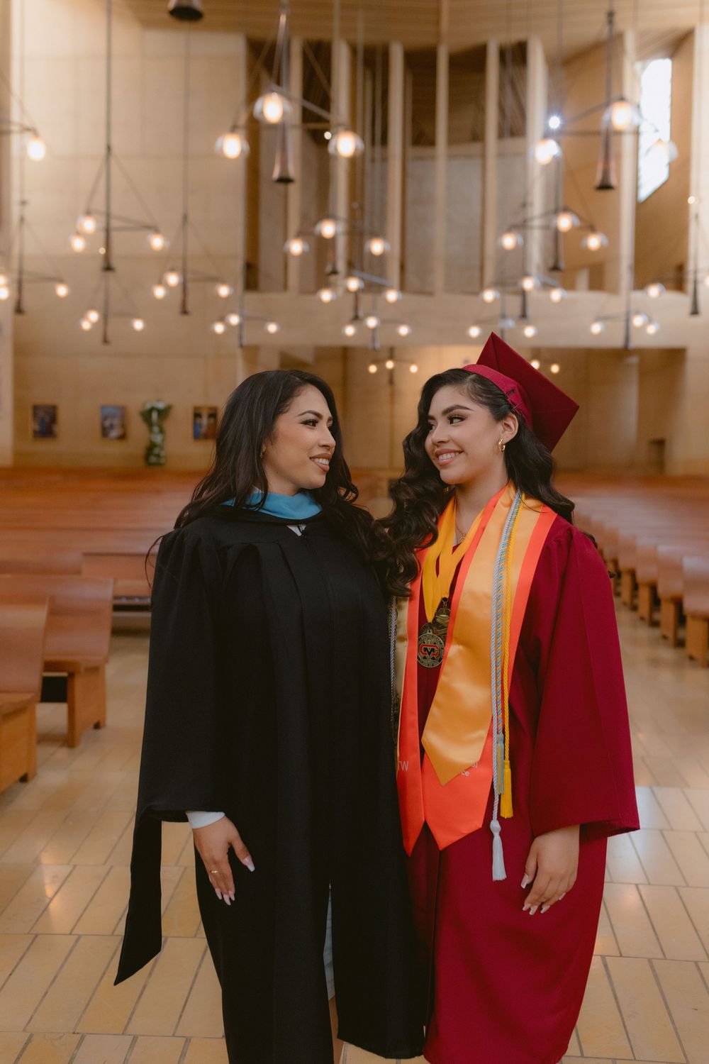 Two graduates in black and red academic regalia pose together inside a church sanctuary with hanging lights.