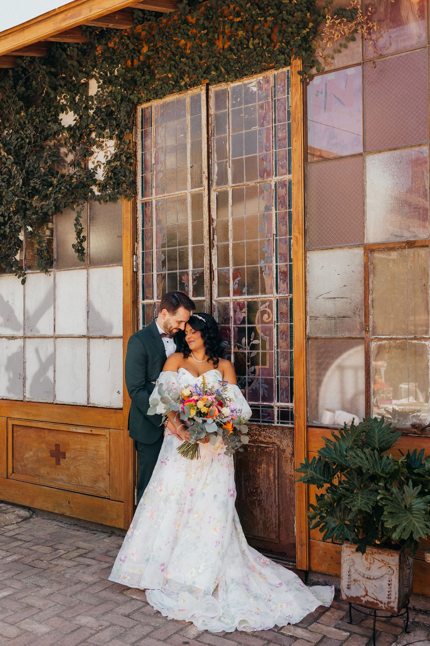 Two people embrace in white bridal attire against a rustic building exterior with stone walls and large windows.