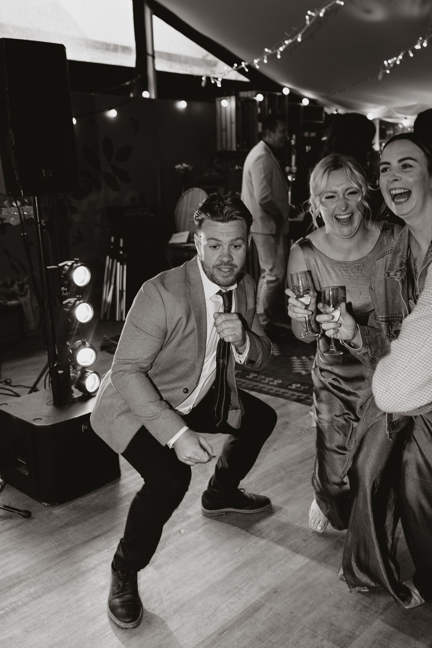 Black and white party photo showing people laughing and celebrating with drinks at a wedding reception on a dance floor.
