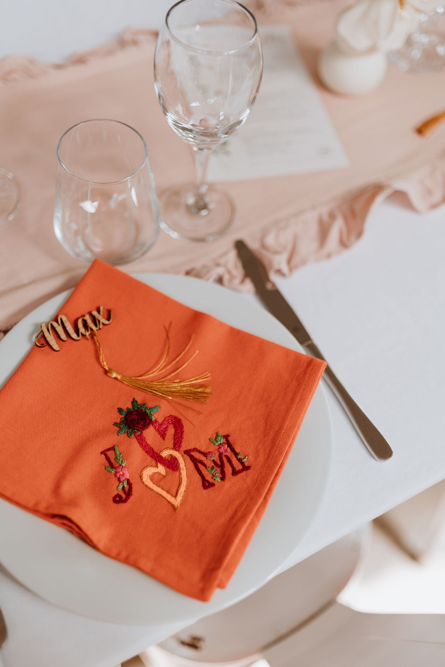 An elegant place setting with an orange napkin embroidered with hearts and initials, wine glasses, and table decor.