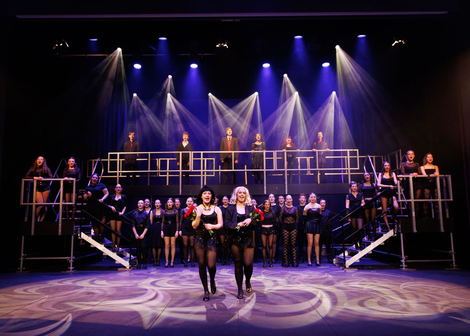 Performers in black outfits sing on a multi-level stage with dramatic blue spotlights and geometric lighting design.