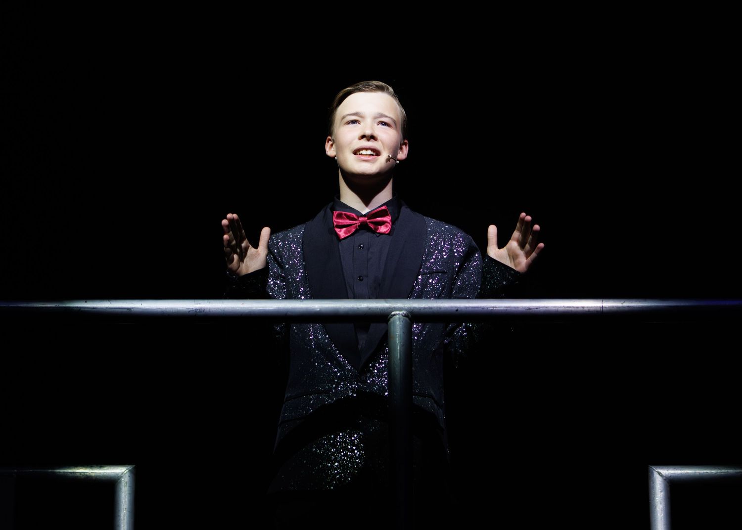 A performer in dramatic lighting gestures while wearing a dark suit and red bowtie against a black background.