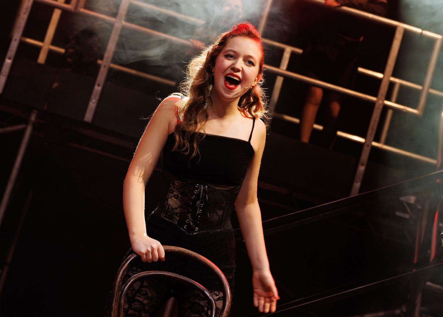 A performer wearing a black dress sings passionately on a dimly lit stage as theatrical smoke swirls in the background.