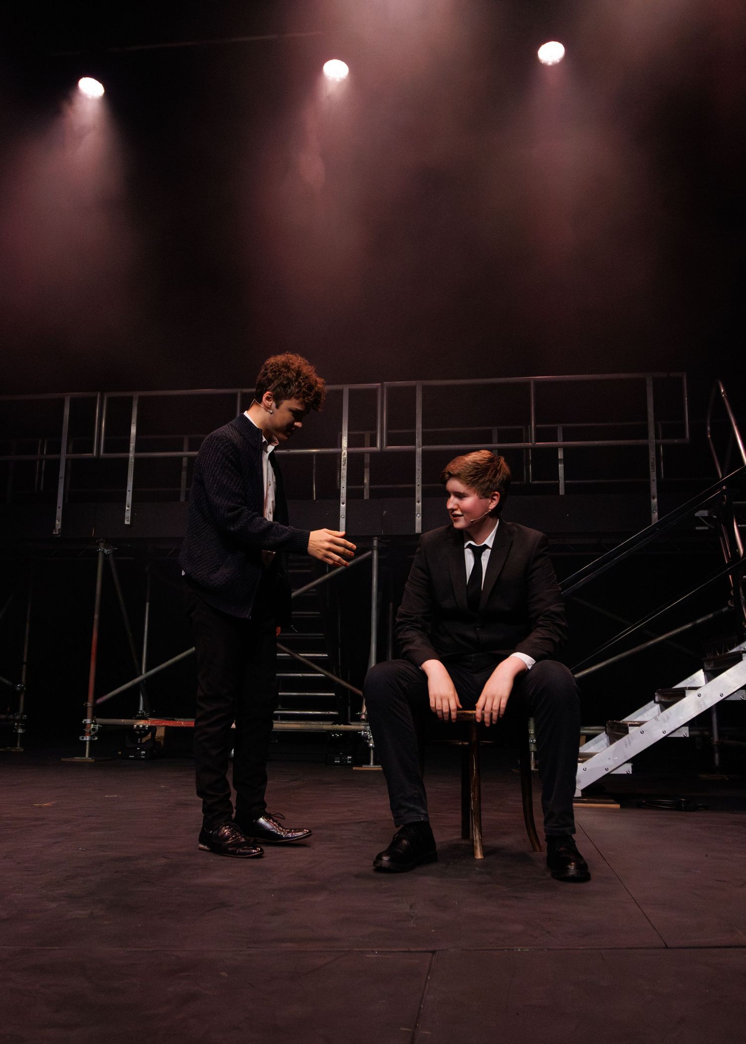Two performers in black suits interact on a dark stage with spotlights and scaffolding in the background.
