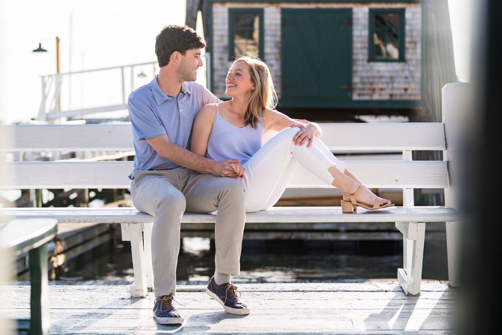 A couple in casual clothing sits together on a white dock by a marina on a sunny day.