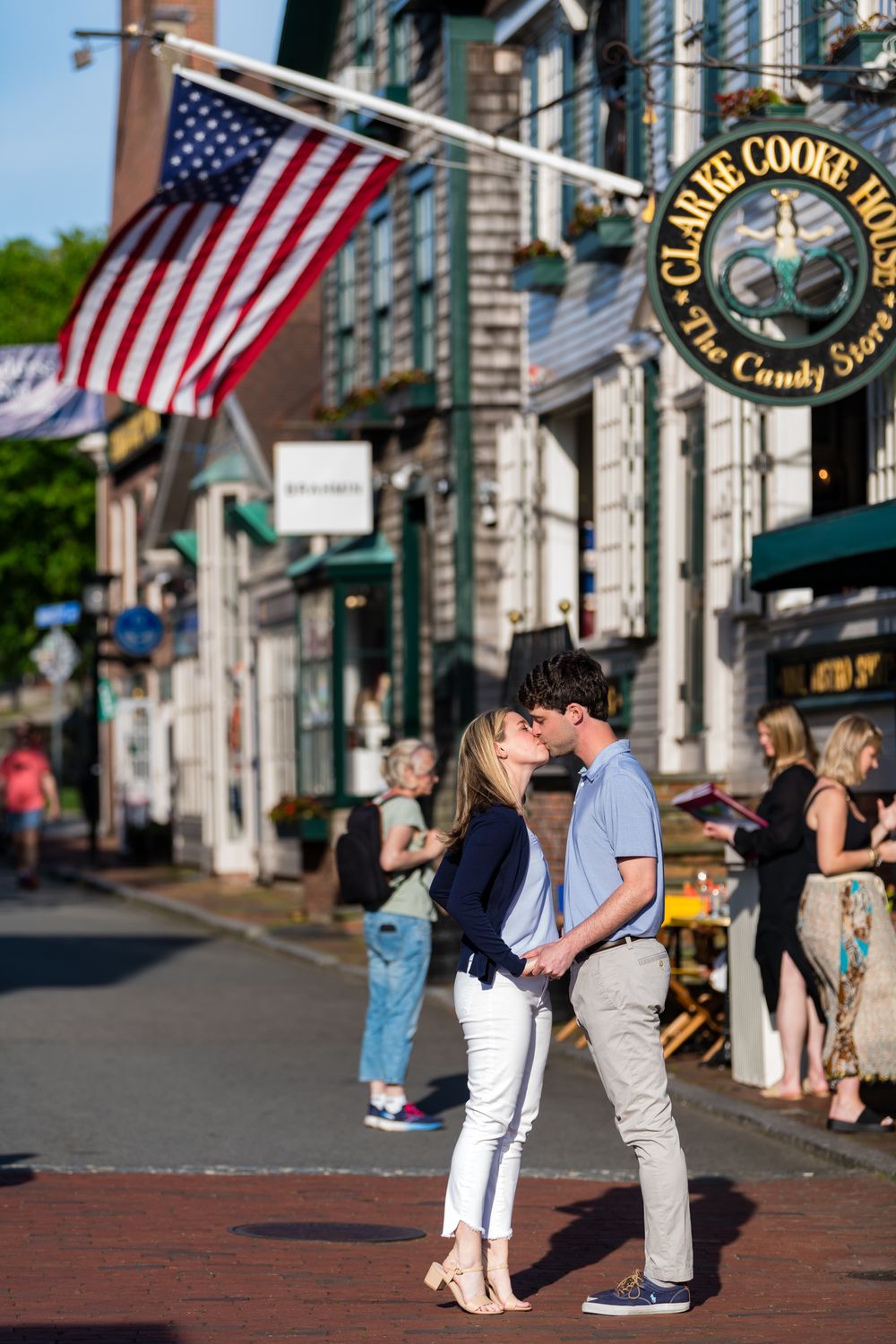 A couple stands on a brick sidewalk beneath an American flag in a quaint downtown shopping district.