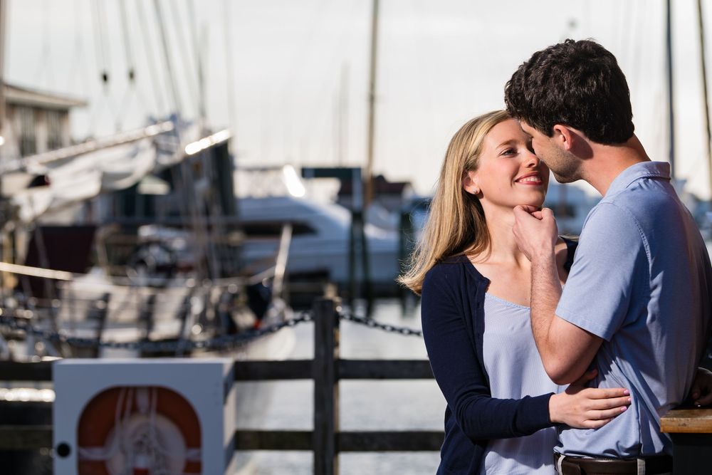 A couple shares a romantic kiss on a boardwalk with boats docked in the marina behind them during sunset.