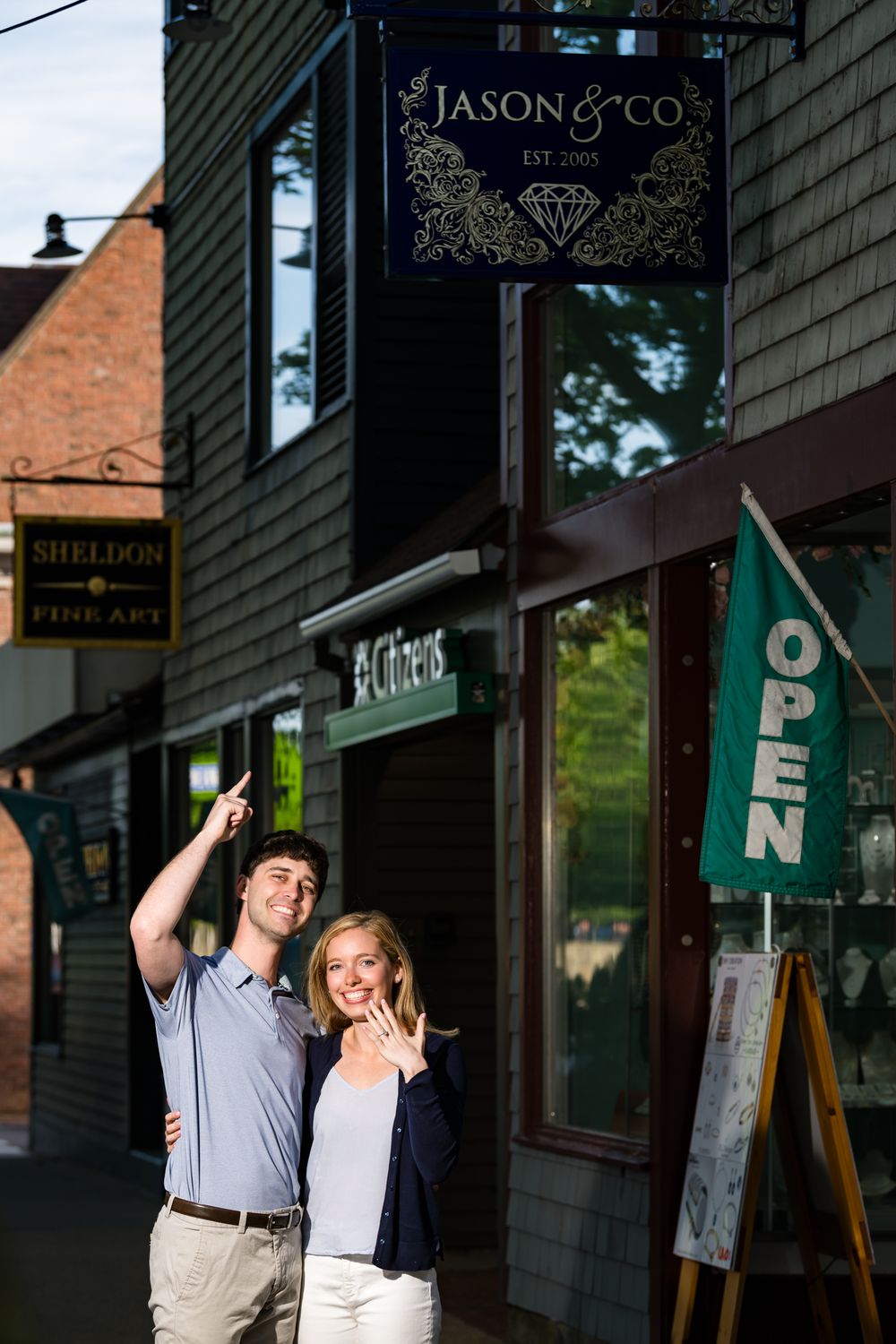 A couple playfully poses in front of storefronts and business signs on a small town main street.