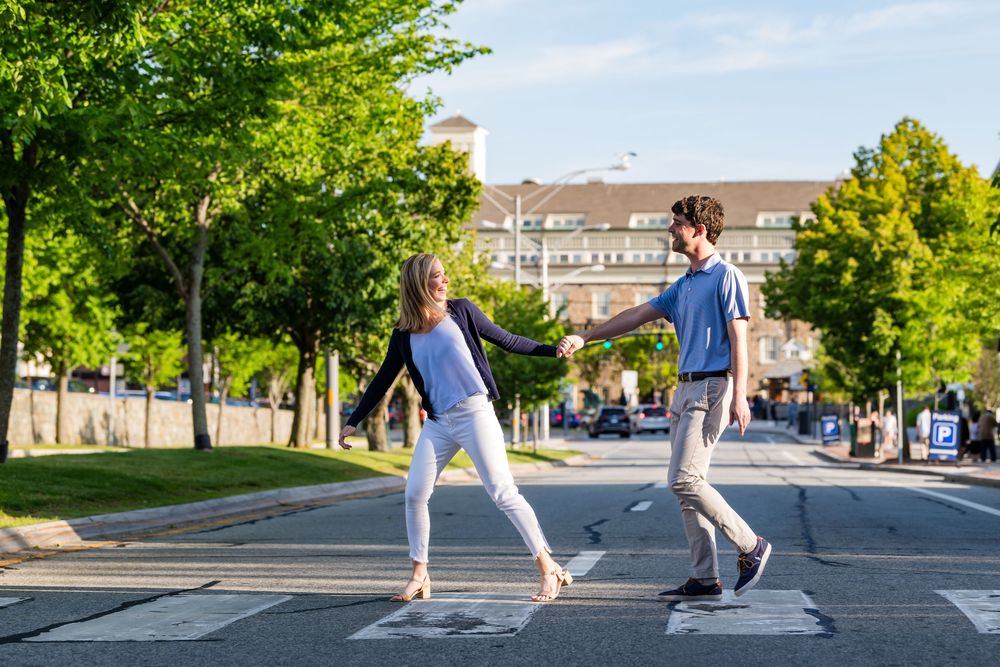 People dancing and leaping in sequence along a sunny sidewalk with trees and buildings in the background.