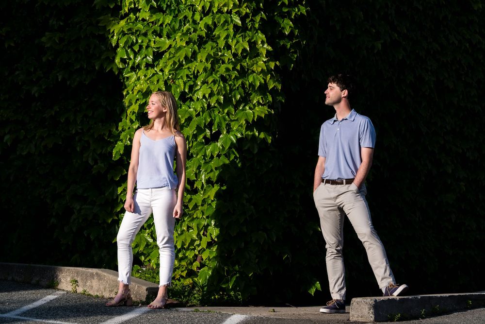 Two people stand against a green ivy wall in casual summer outfits on a sunny day.