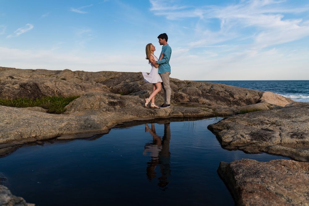 A romantic couple stands reflected in a tidal pool on rocky coastal terrain at sunset.