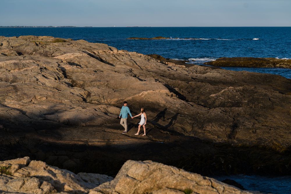 Two figures walk hand in hand across rocky coastal terrain with blue ocean waves and horizon in background.