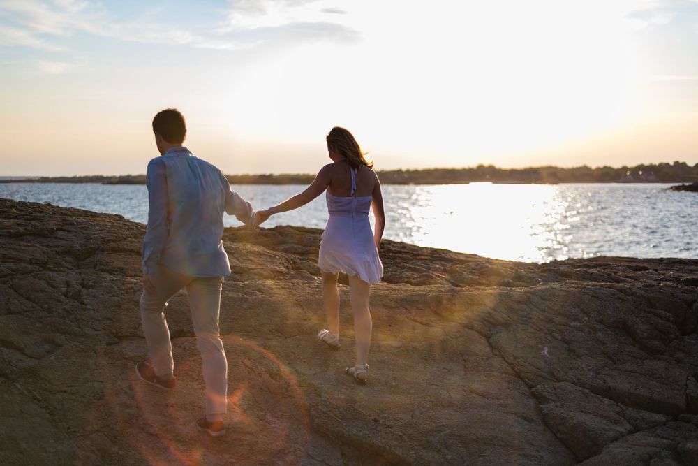 Silhouettes of two people walking along a rocky coastline at sunset with golden light reflecting on the water.