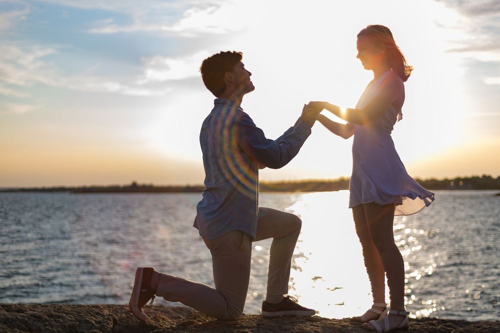 A romantic proposal scene silhouetted against a golden sunset over the water.