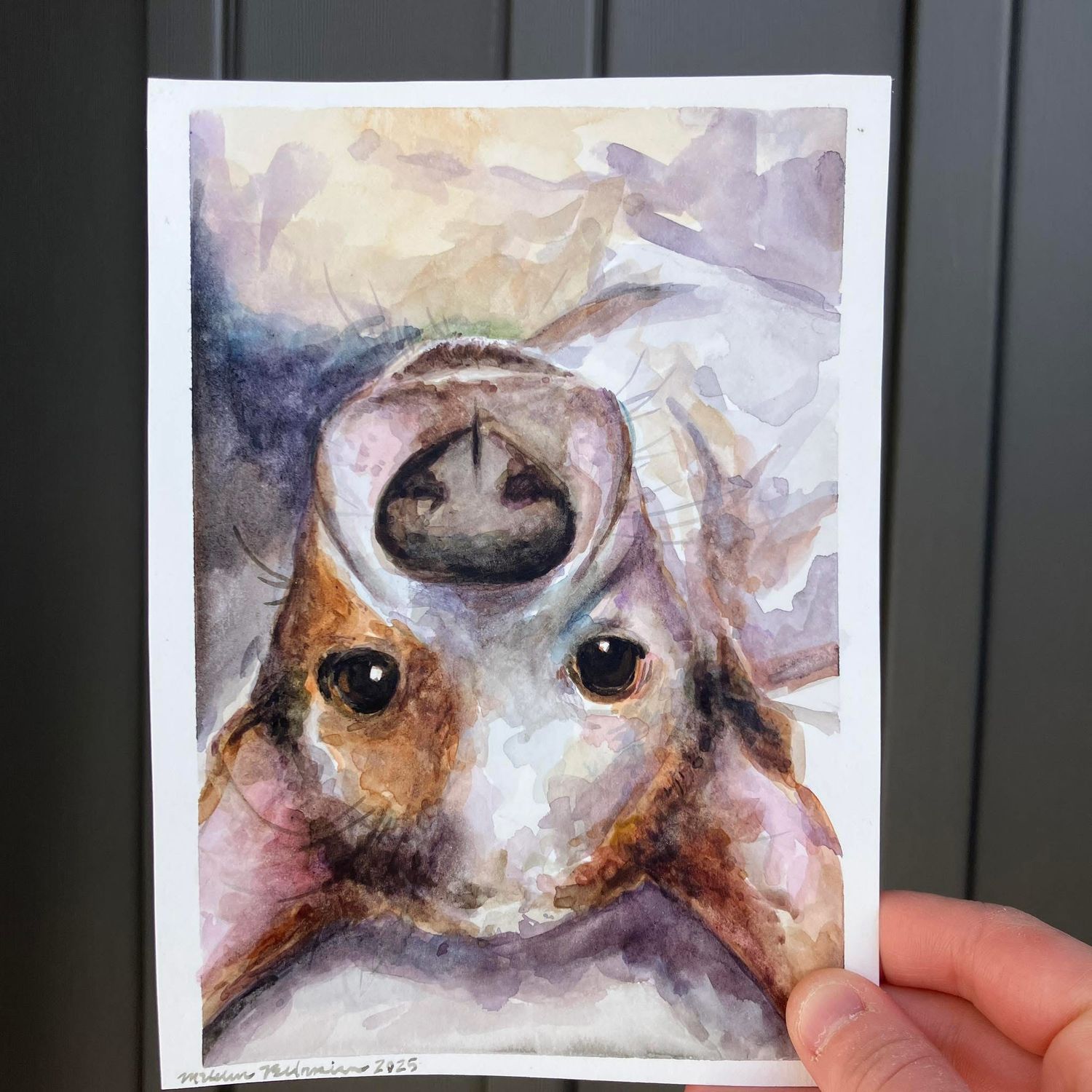 A whimsical watercolor painting of a dog lying on its back, gazing up with expressive eyes, rendered in soft, warm tones and held in hand