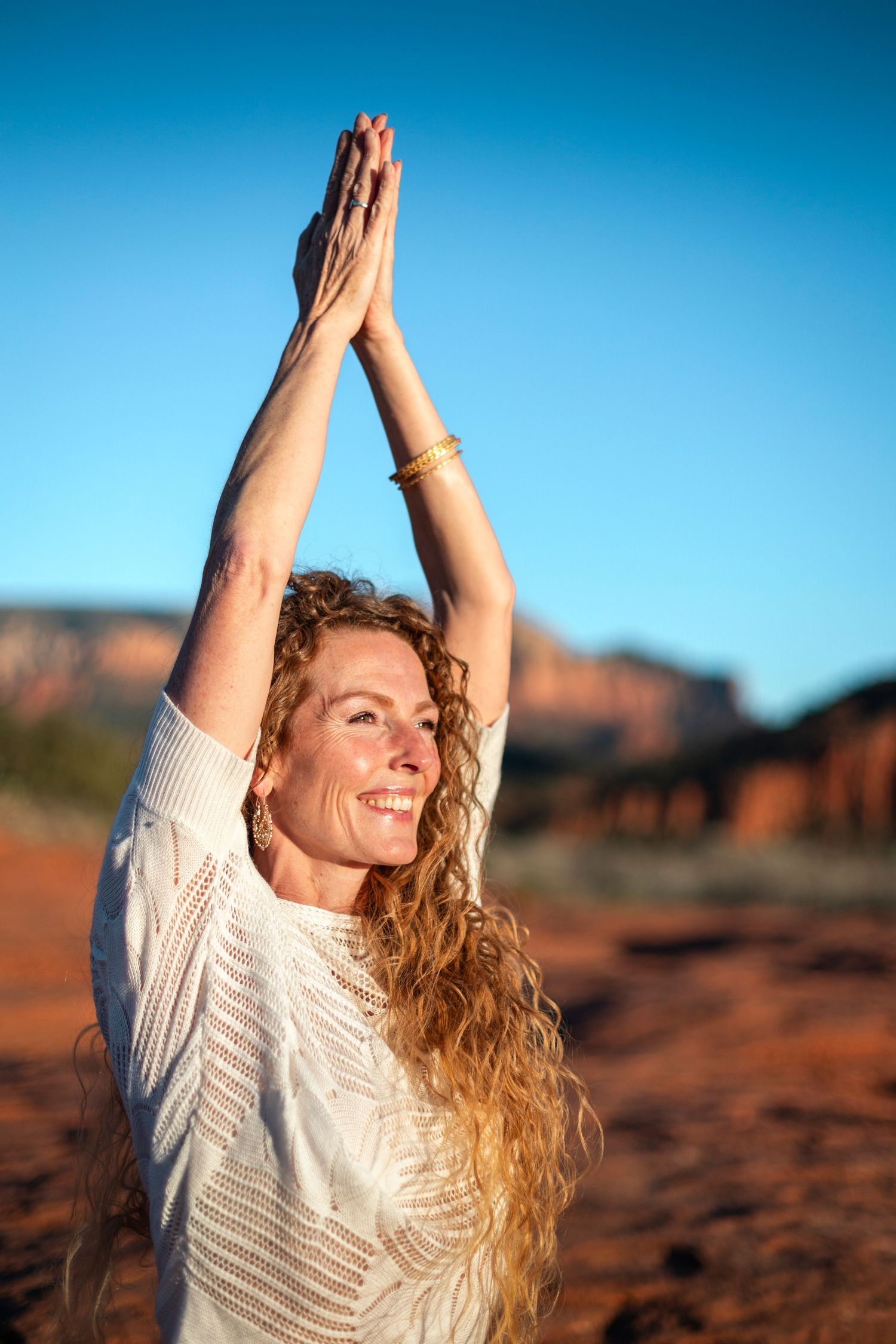 Smiling blonde woman in a light green maxi dress poses on a rocky beach with palm trees at sunset.