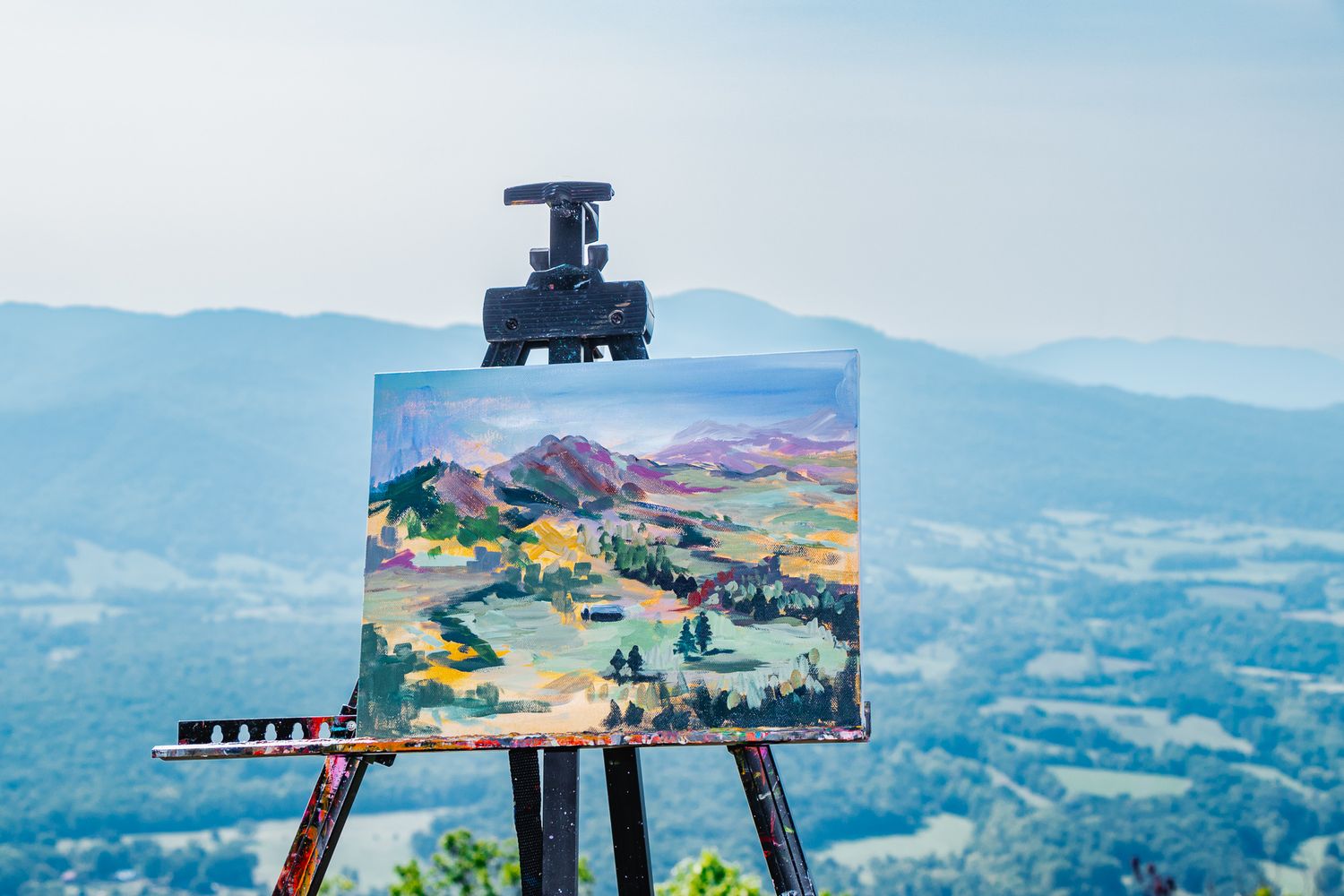 A colorful landscape painting on an easel, depicting rolling hills and valleys, set against the matching backdrop of a Blue Ridge Mountain