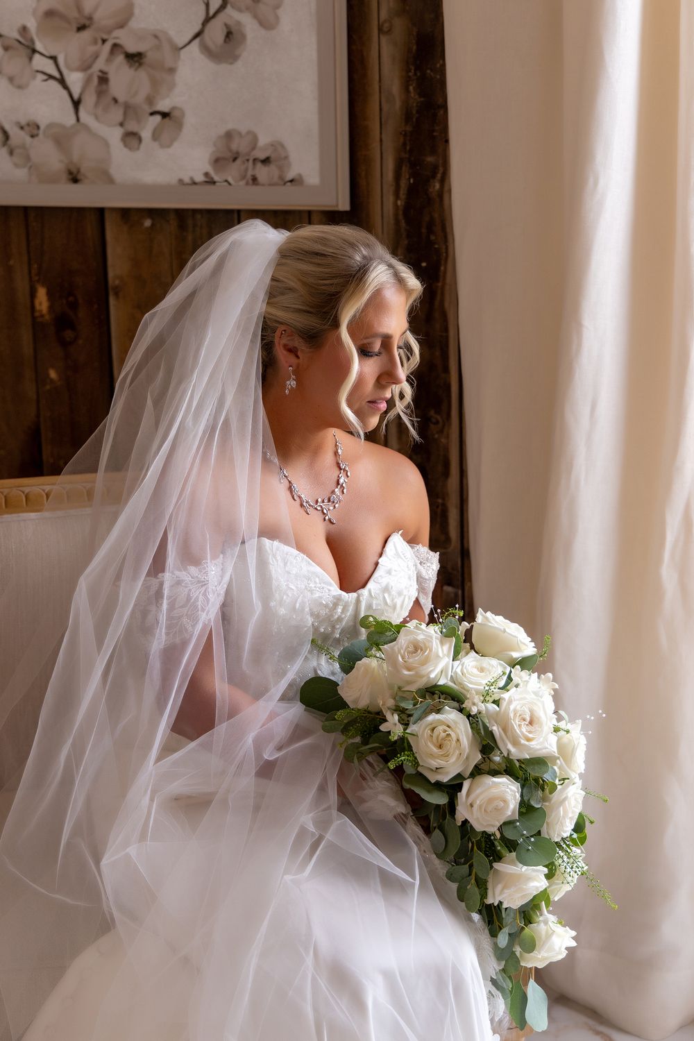 Bride in a white wedding dress and flowing veil holds a bouquet of white roses against natural window light. Wedding photo Bicentennial Barn