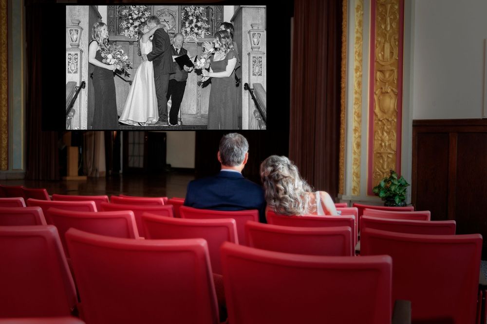 Two people watch a vintage wedding scene projected on a movie screen in a theater with red seats. Wedding photo Engineer Club Dayton Ohio.