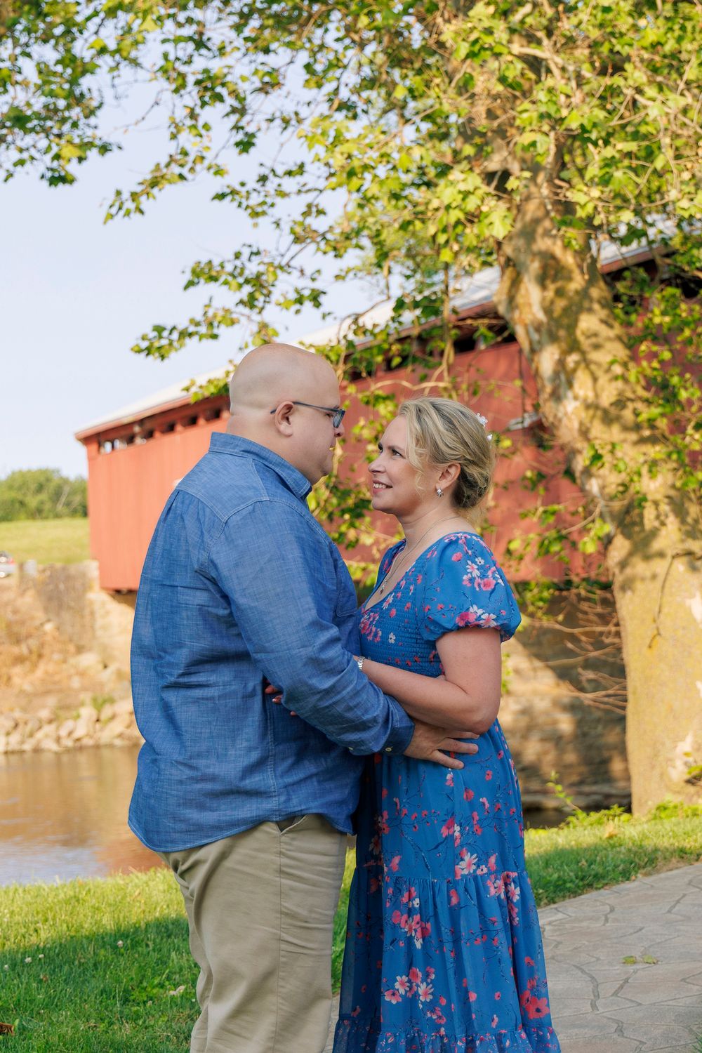 Couple shares a romantic moment by a historic red covered bridge on a sunny day in a scenic outdoor setting. Engagement Session Troy Ohio.