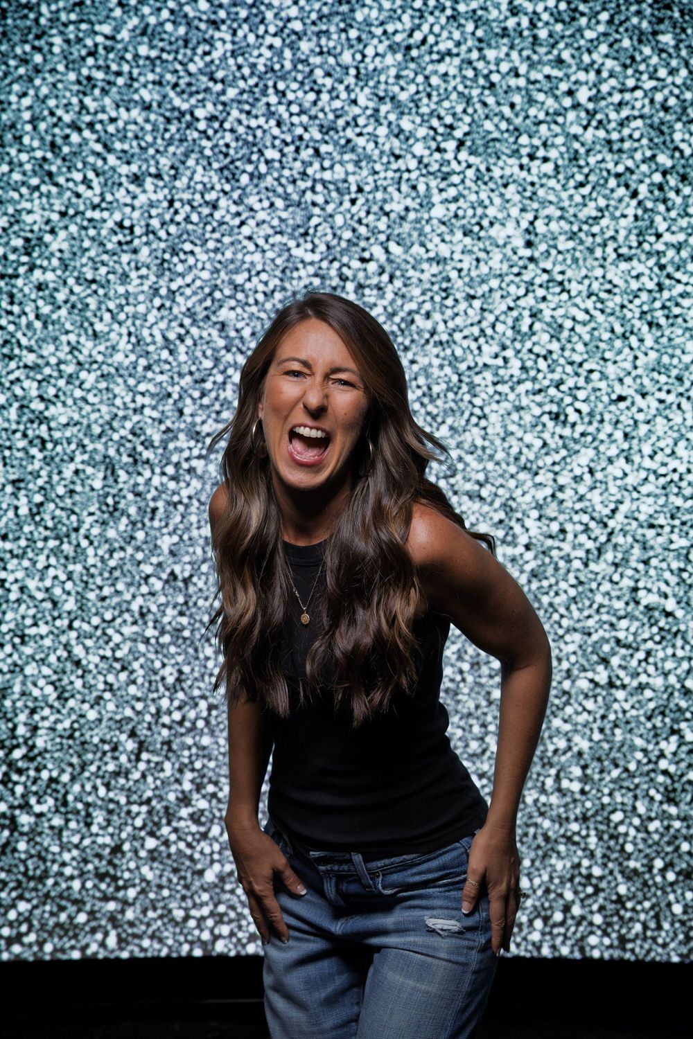Energetic woman in black tank top and jeans laughs expressively against a sparkly blue backdrop.