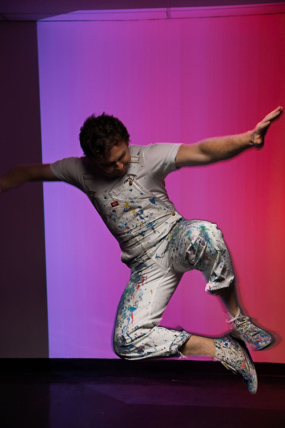Dancer leaping in white paint-splattered outfit against vibrant purple and red gradient background.