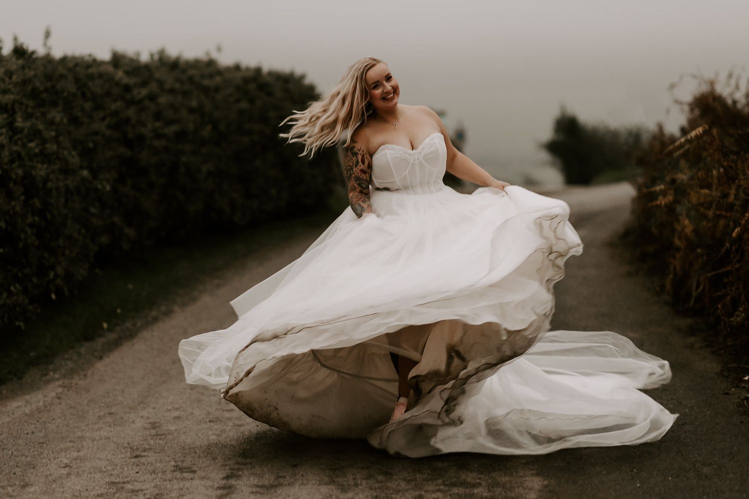 A bride in a flowing white wedding dress twirls on a dirt path surrounded by greenery at dusk.