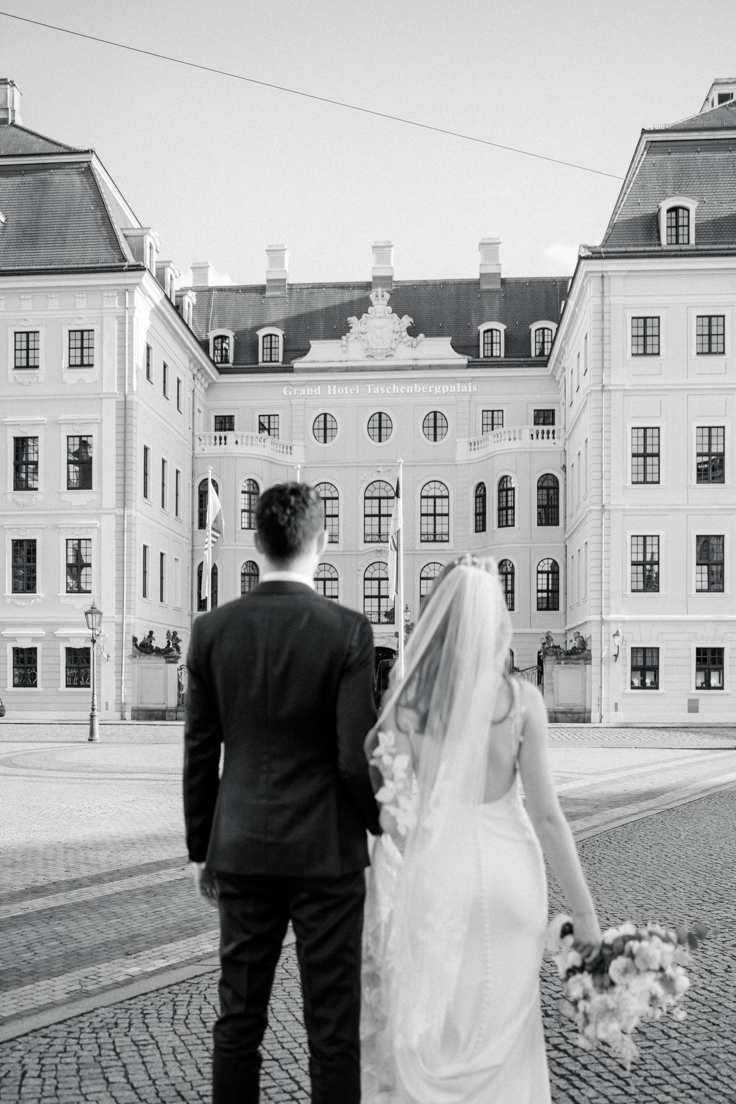 A couple in wedding attire views a grand historic European palace building with classical architecture.