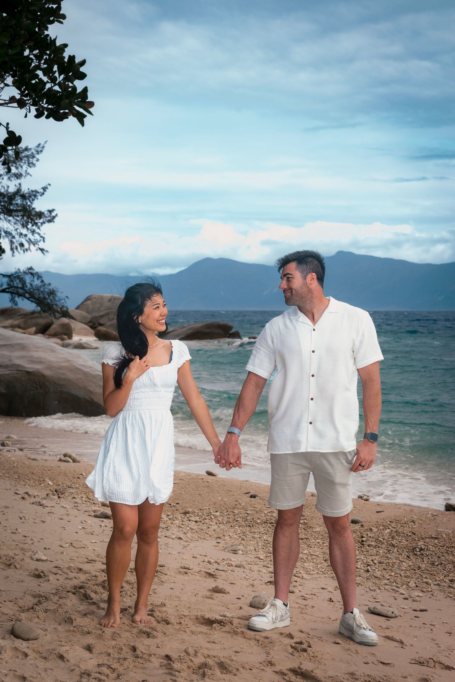 Couple in white outfits holding hands and laughing while standing on a tropical beach with mountains in the background.