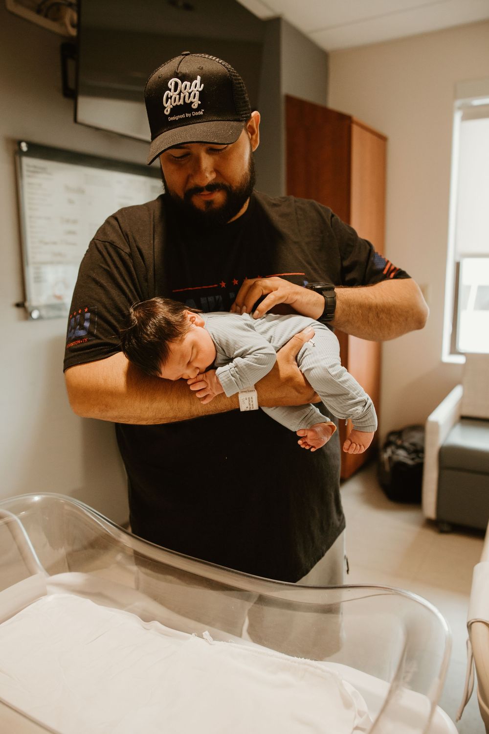 A person in a black shirt and baseball cap tenderly holds a sleeping newborn baby in a hospital room.