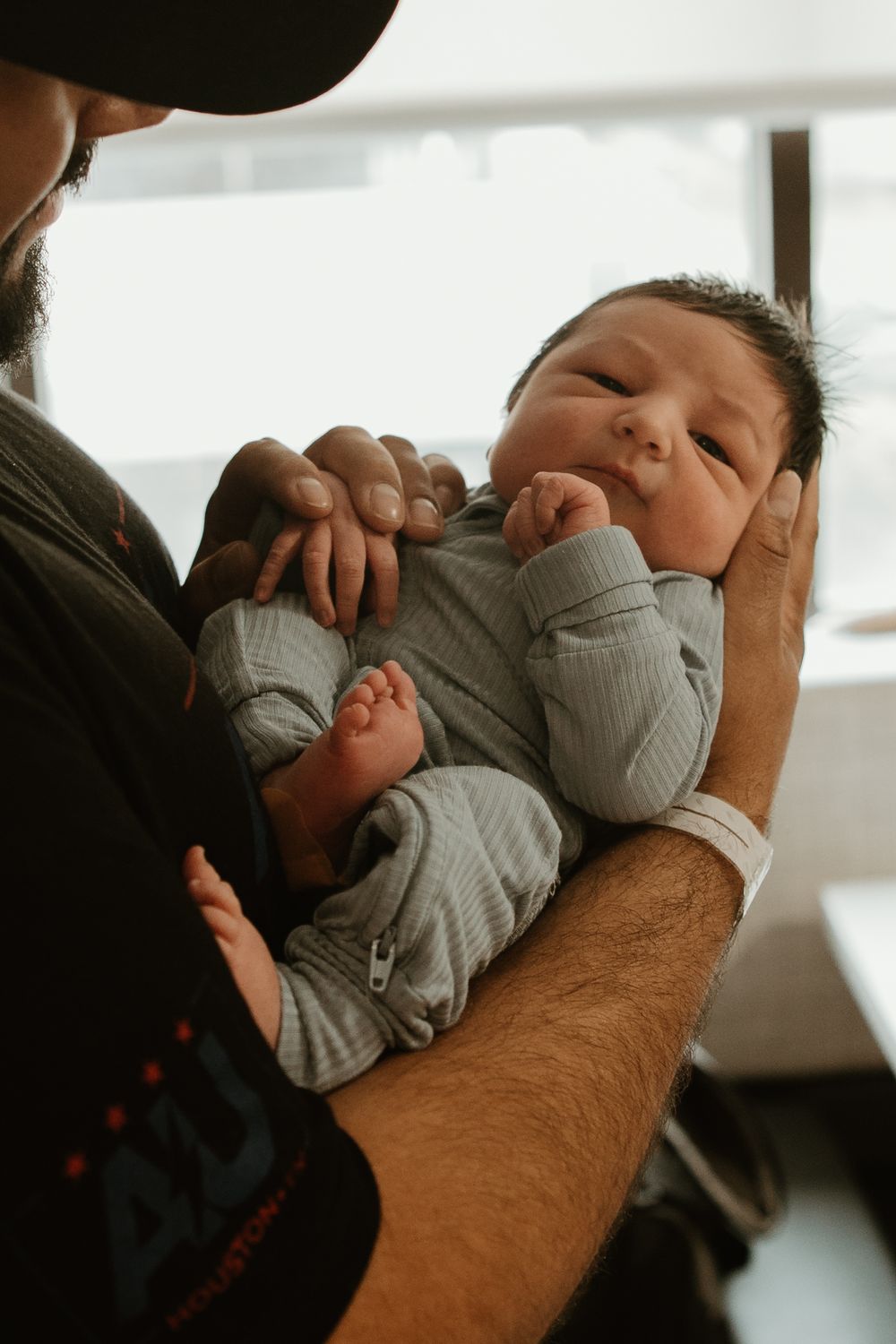 Tender moment of a baby in light gray onesie being held in strong arms against warm lighting.