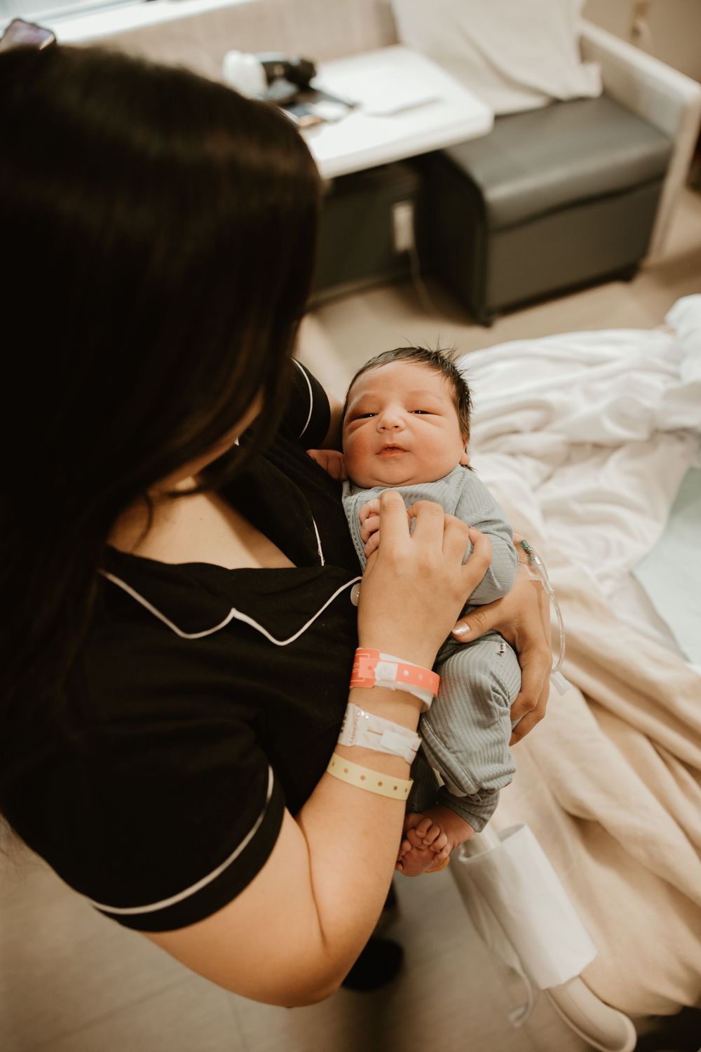 A newborn baby in a hospital room rests on a bed while wearing a light blue onesie, with hospital ID bracelets visible.