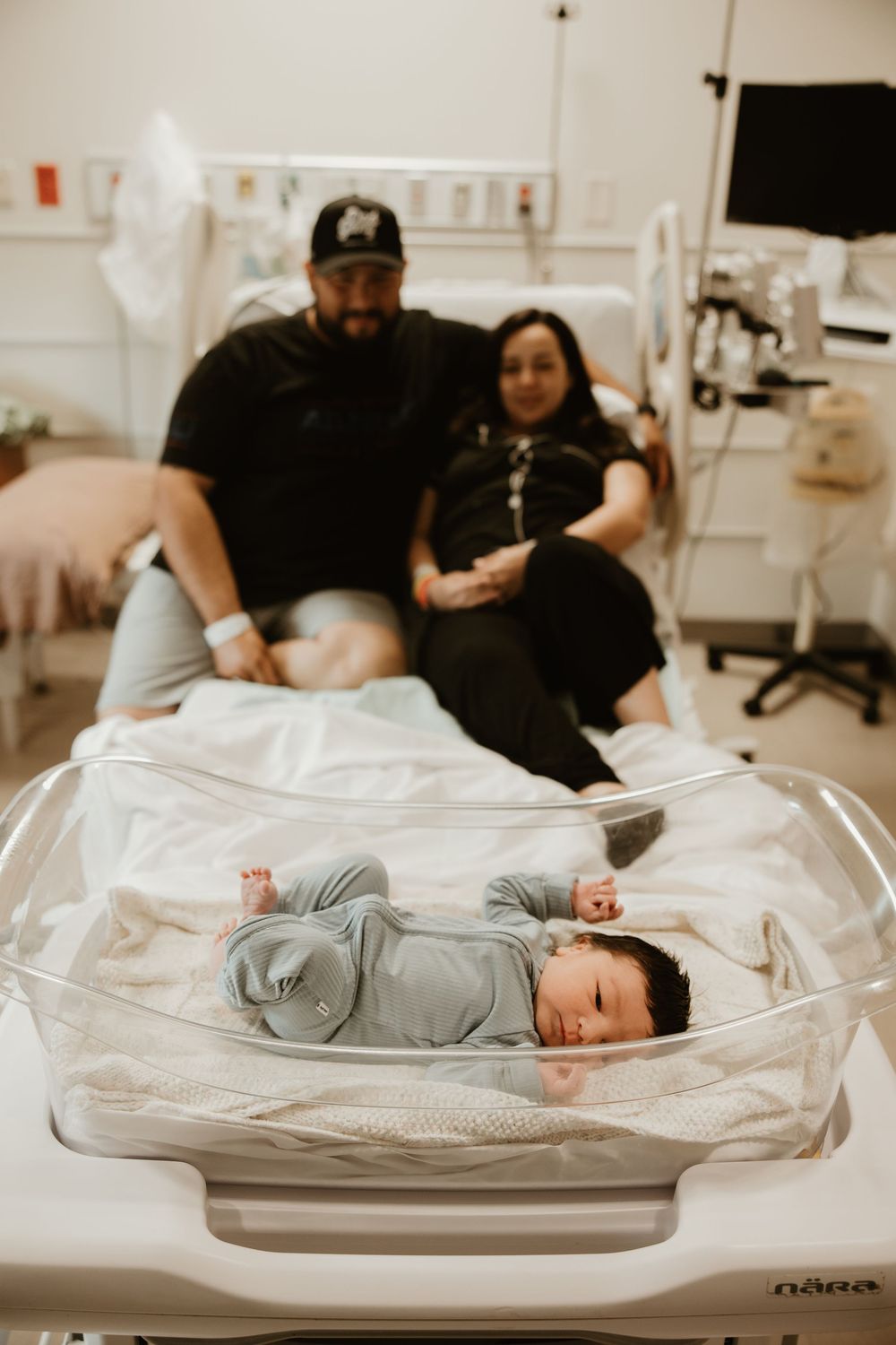 A sleeping newborn in gray clothing lies in a hospital bassinet while two adults watch from the nearby bed.