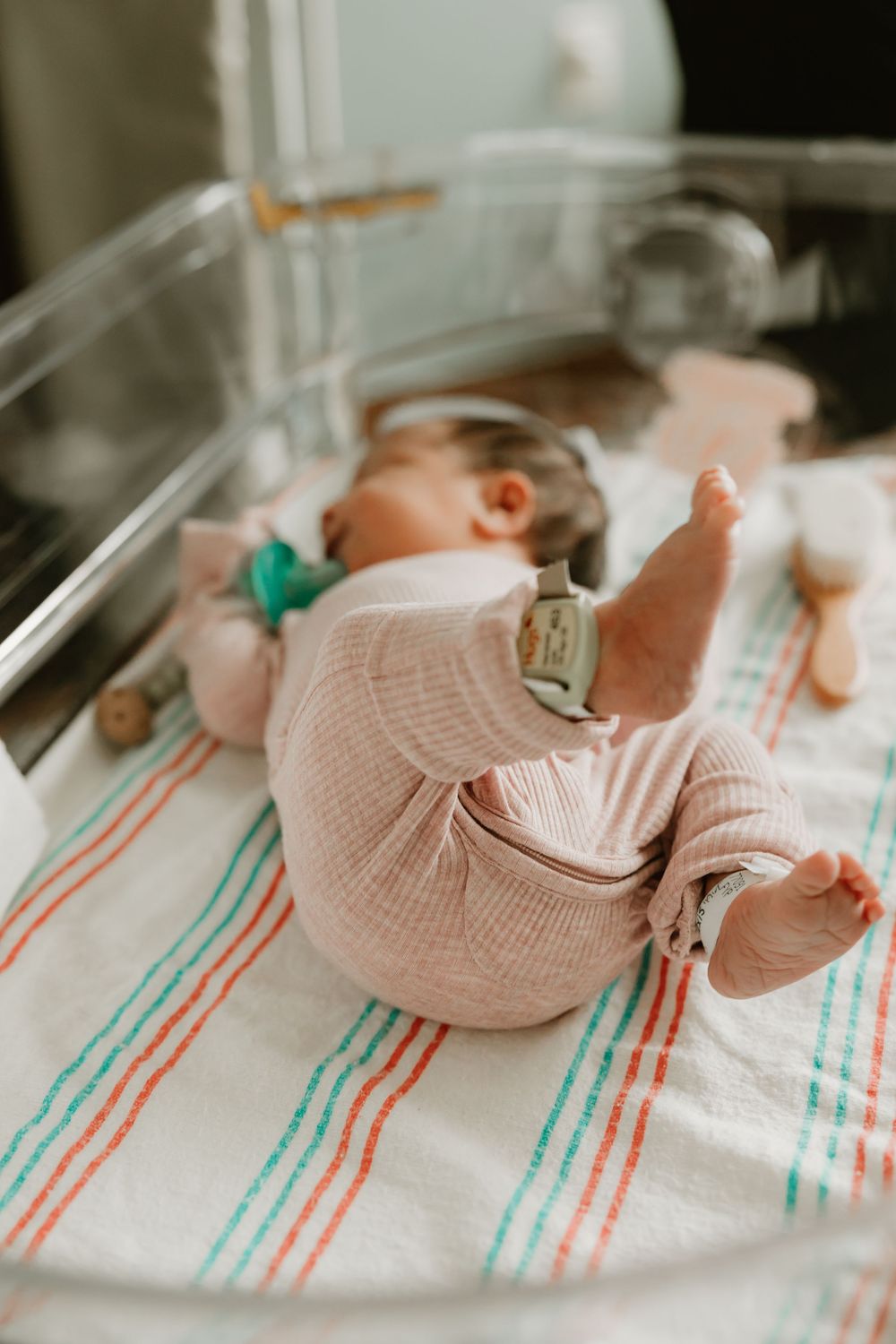 Premature baby sleeps peacefully in hospital incubator with monitoring devices and striped blanket.