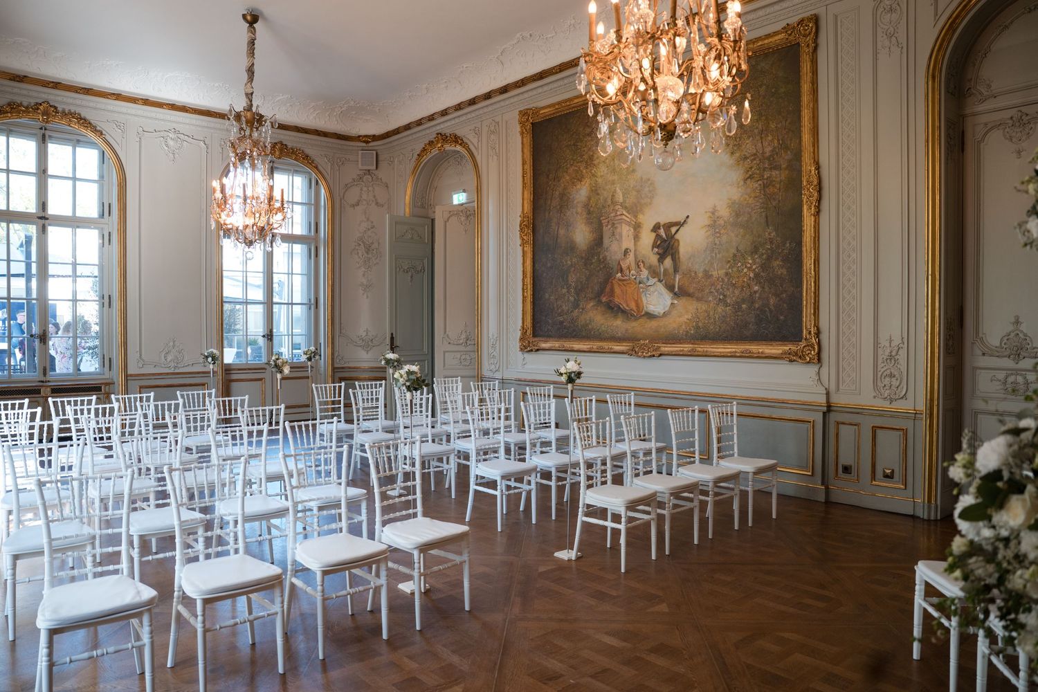 Elegant ballroom with crystal chandeliers, white chairs and classical artwork features ornate French-style architectural details.