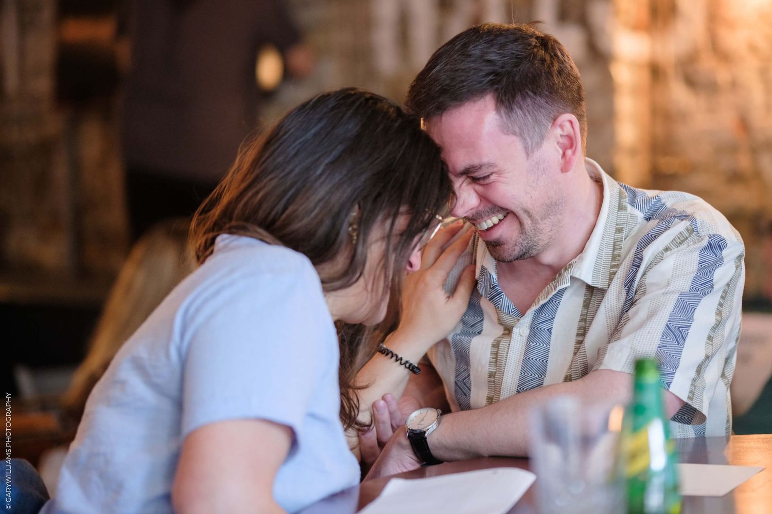 A couple shares an intimate laughing moment together at a table in a restaurant setting.