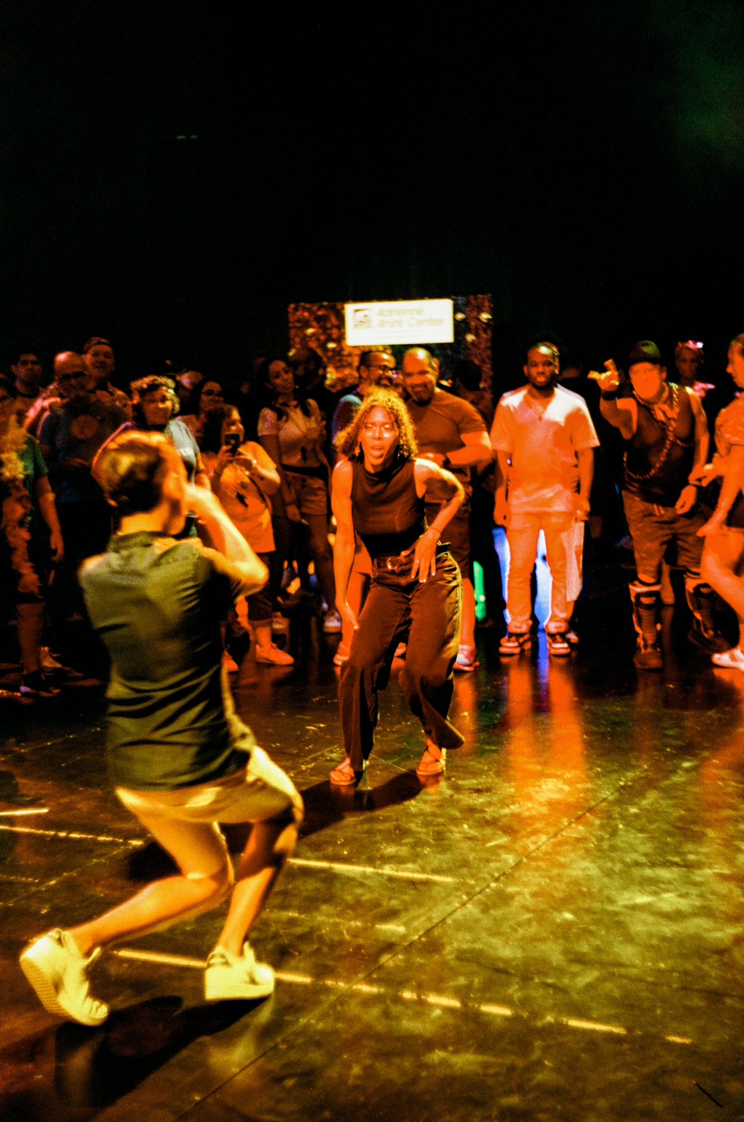 Dancers perform energetic swing dance moves in a dimly lit ballroom with warm orange lighting and a crowd watching.