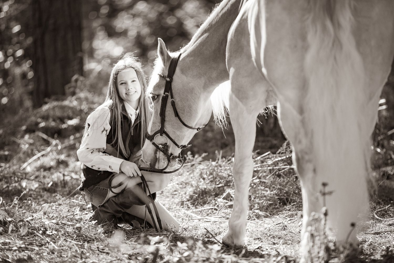 A person in western attire kneels beside a majestic white horse in a black and white forest setting.