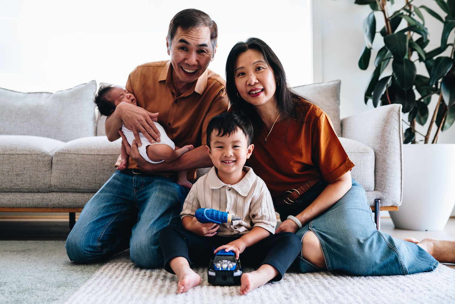 A joyful Asian family plays together and laughs while relaxing on a gray couch at home taken by melbourne family photographer.