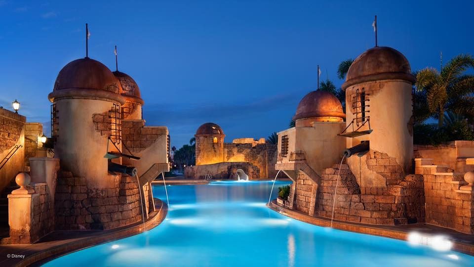 Illuminated swimming pool surrounded by castle-like turrets and stone architecture at Disney's Caribbean Beach Resort at dusk.