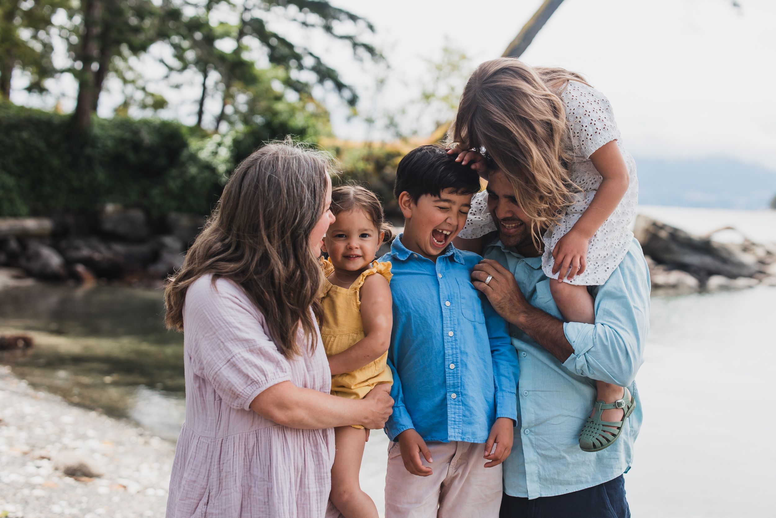 Family enjoying a candid moment and laughing together on a rocky beach with mountains visible in the background.