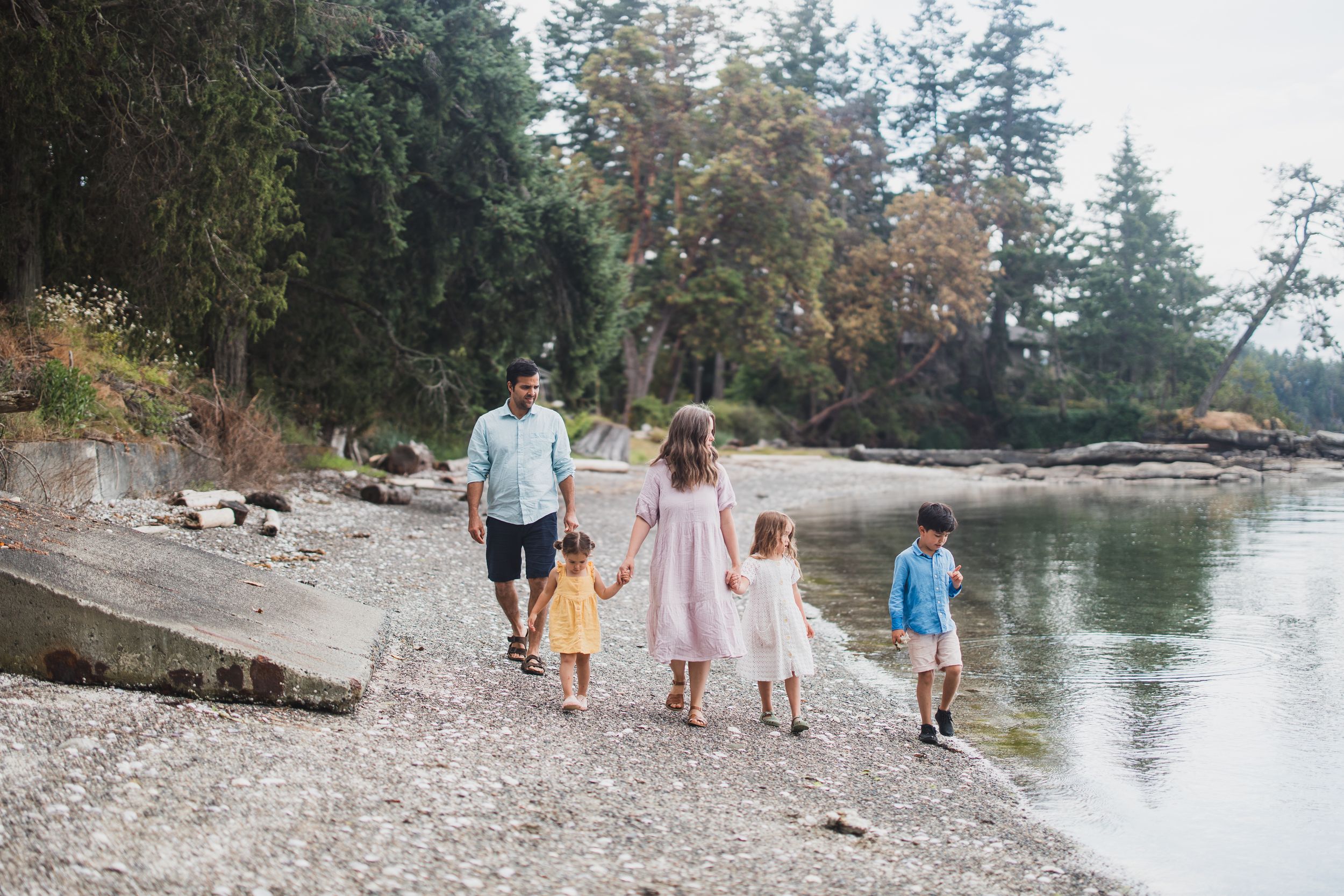 Family strolls along scenic rocky beach shoreline with evergreen trees and calm waters on a peaceful autumn day.