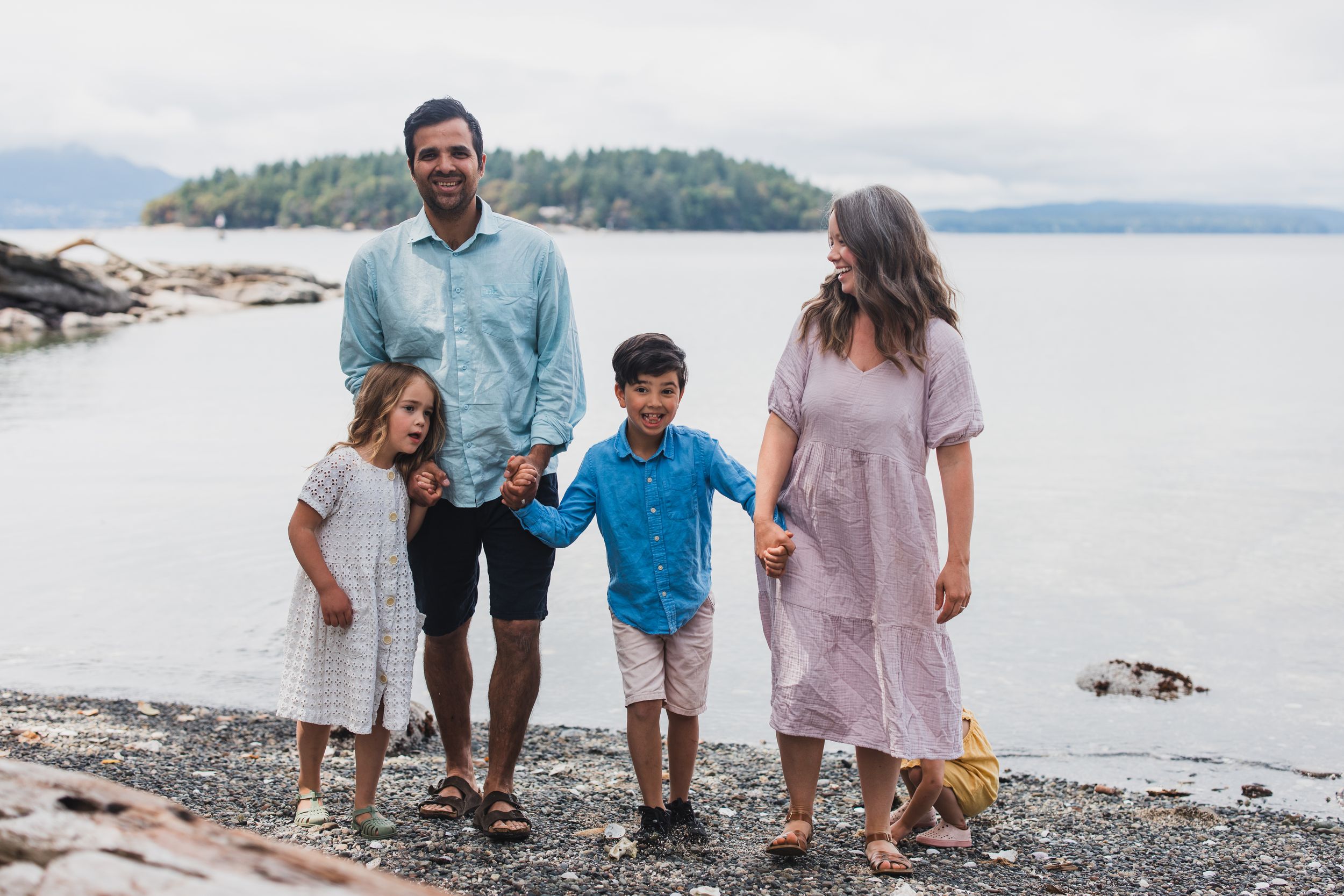 A family walks together along a rocky beach shoreline with mountains and water in the background on a cloudy day.