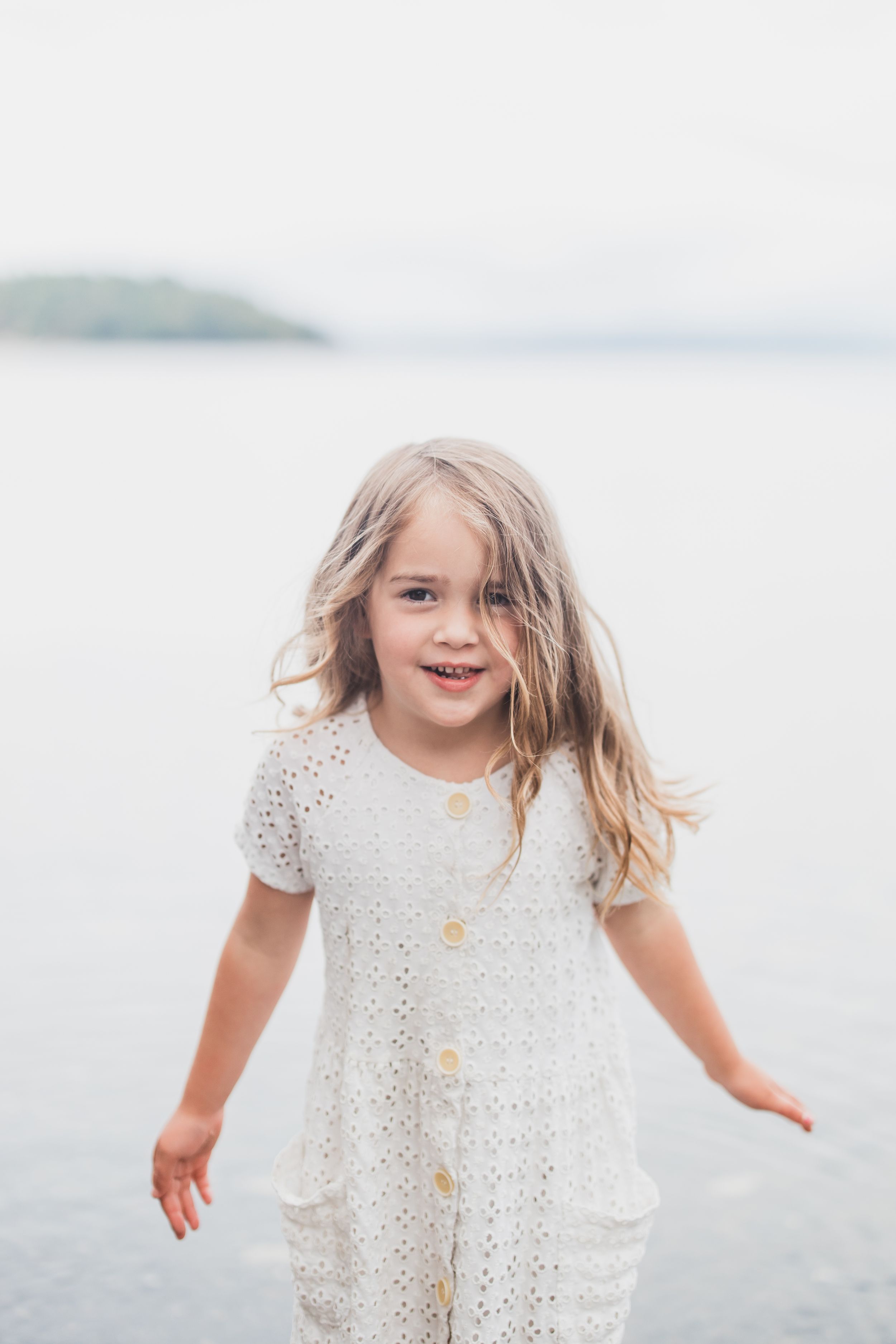Young girl in white lace dress laughing and playing by the waterfront on a cloudy day.