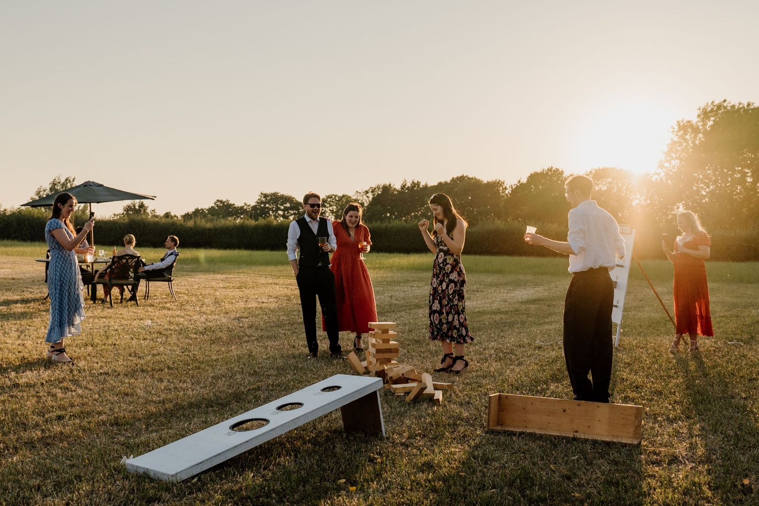 Guests playing cornhole lawn game at golden hour during an outdoor wedding reception on a sunny day.