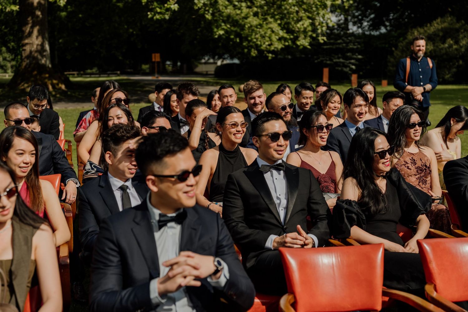 Large crowd of formally dressed wedding guests gathered at sunny outdoor venue.