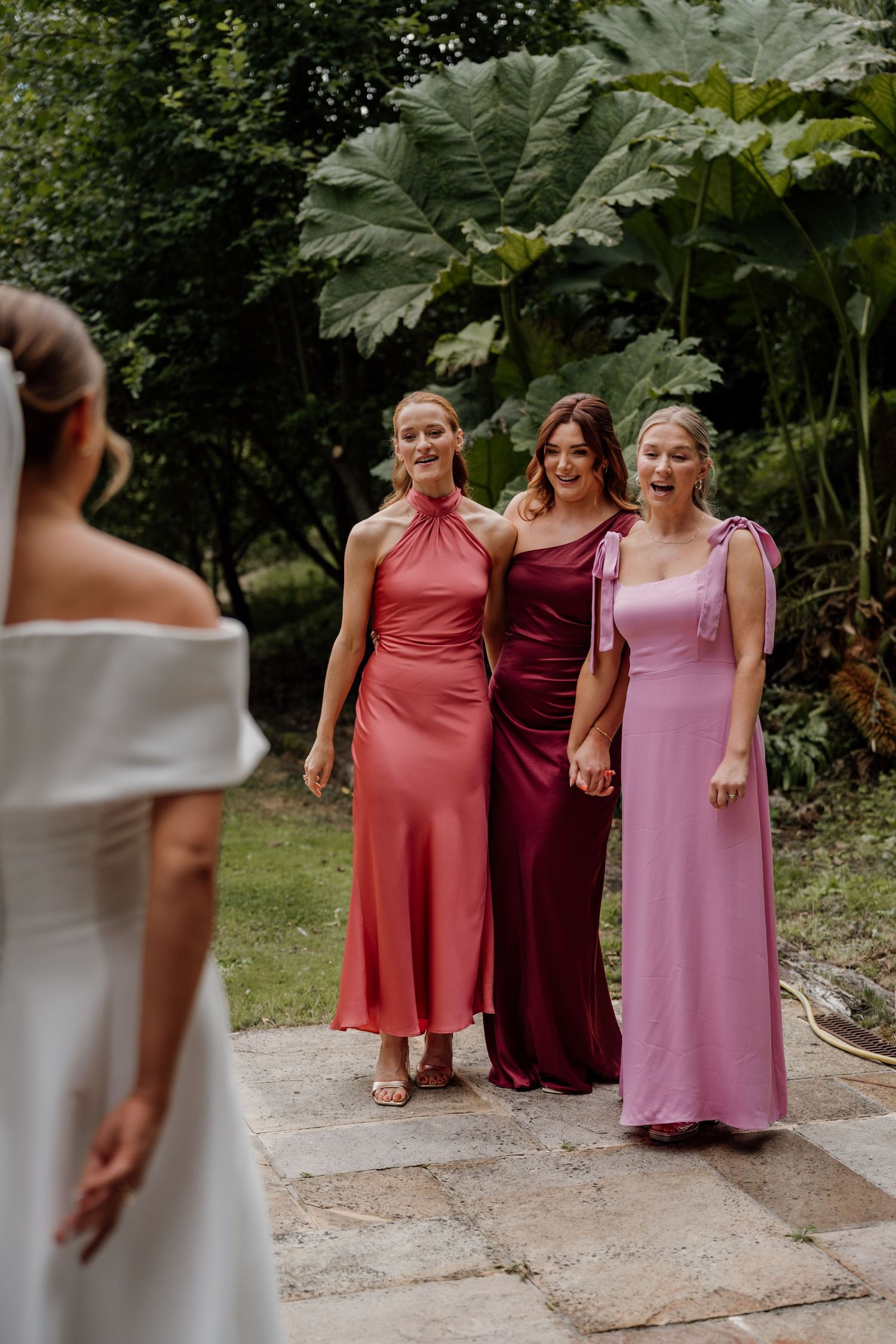 Bridesmaids in pink and burgundy dresses pose together on a garden path during a wedding celebration.
