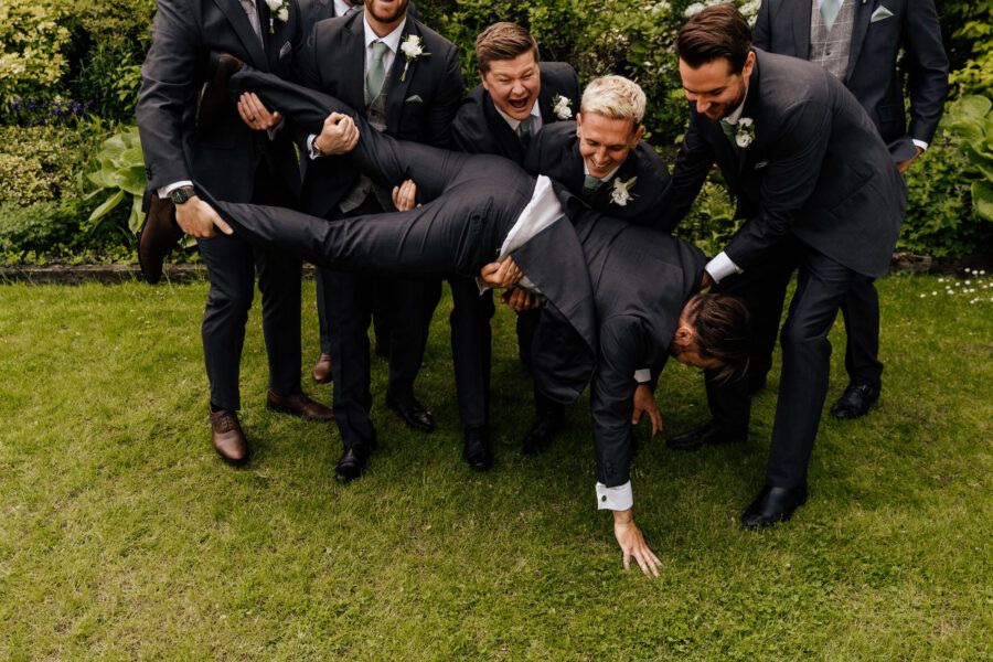 Group photo of groomsmen in dark suits lounging casually on grass during an outdoor wedding.